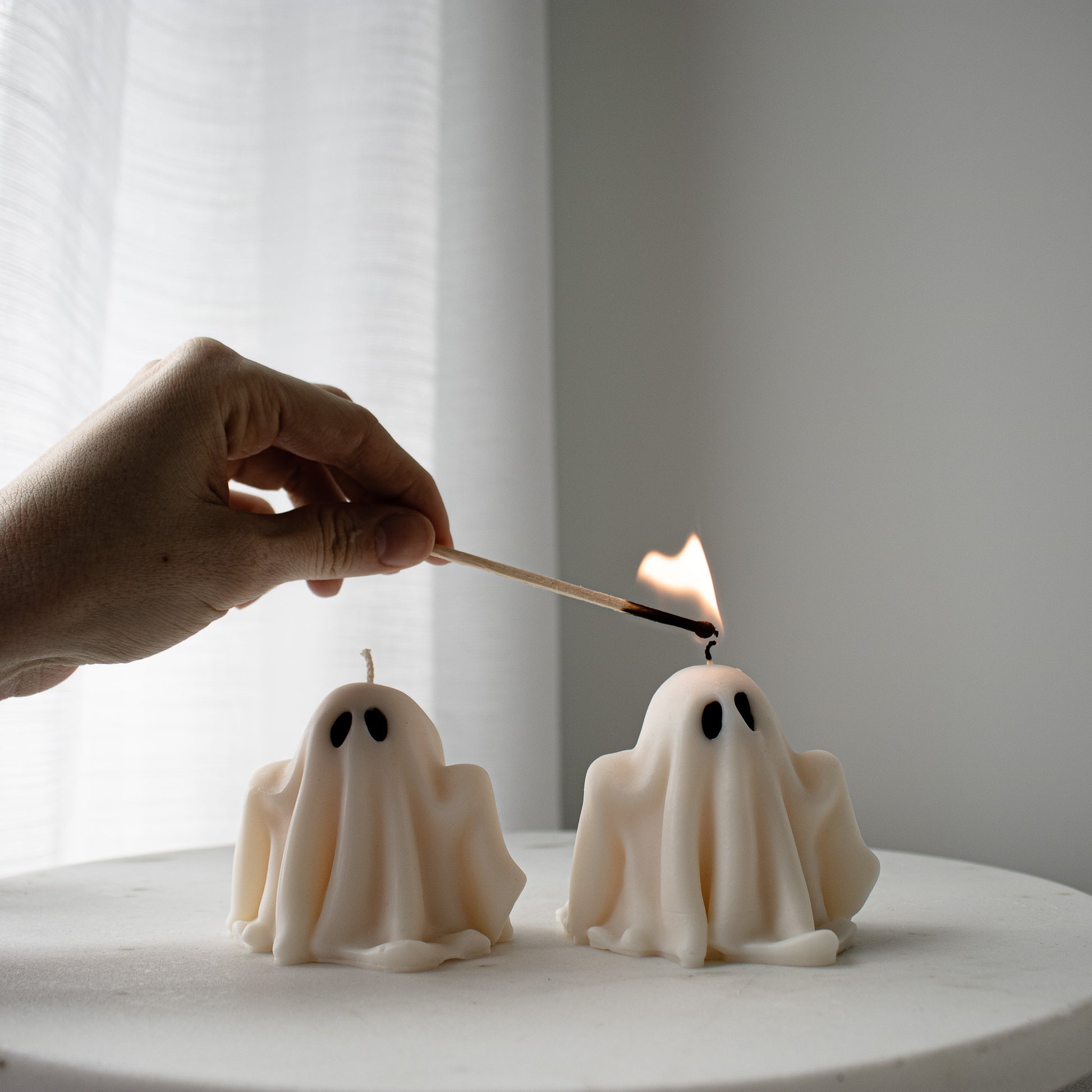 Person lighting a match to a ghost-shaped candle on a white surface with a light background