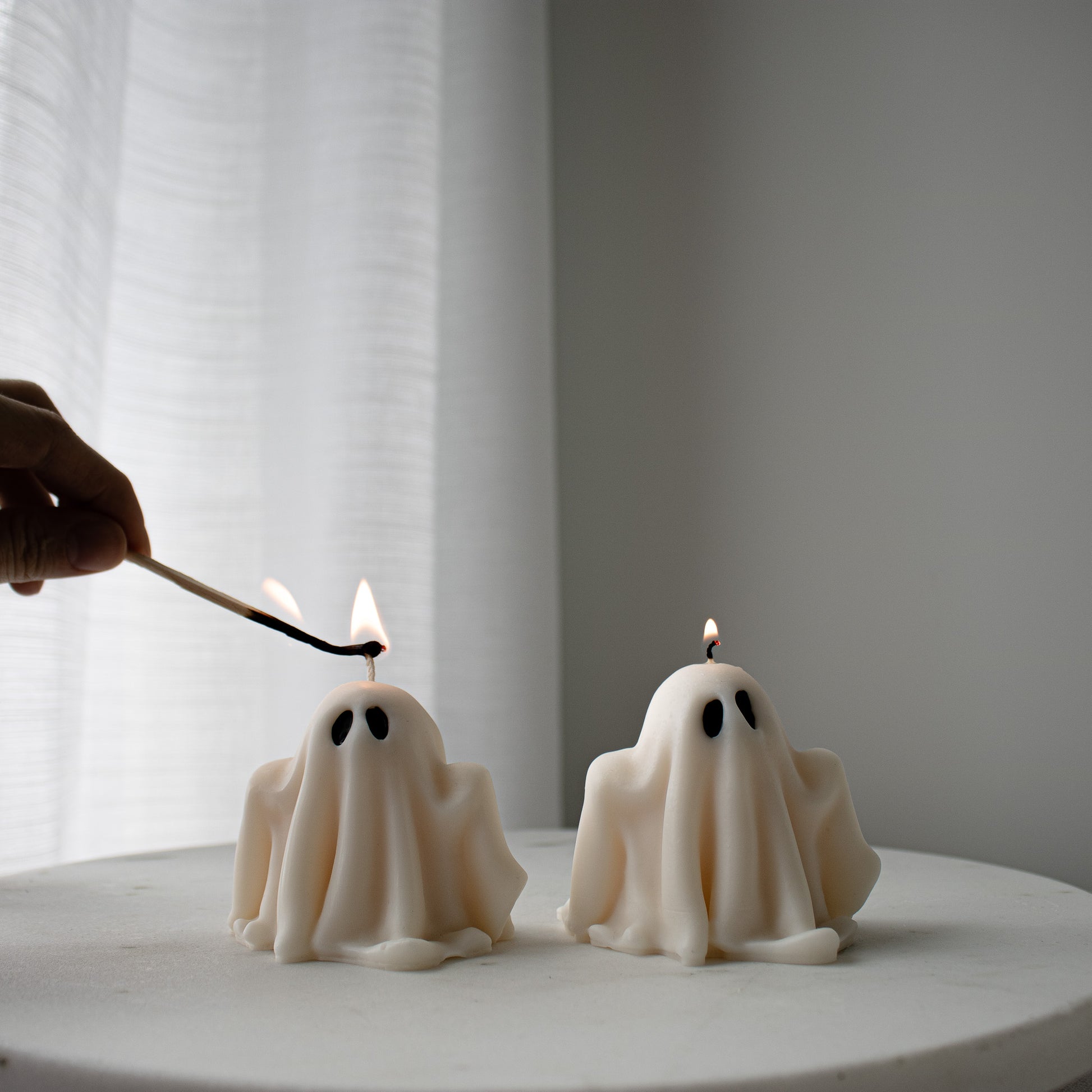 Two ghost-shaped candles being lit with a match on a white surface.