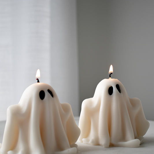 Two ghost-shaped candles with lit wicks on a neutral background